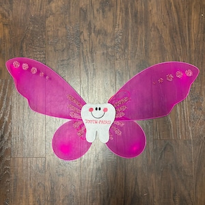 May include: A pair of bright pink sheer fairy wings featuring a central white felt tooth applique with a smiling face and the text "TOOTH FAIRY" embroidered in pink. The wings are accented with pink glittery details and small circular embellishments.