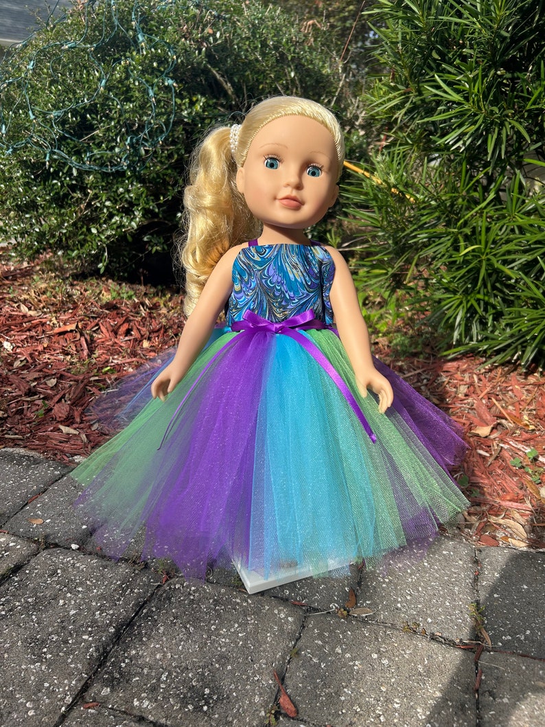 May include: A doll wearing a blue and green patterned dress with a purple tulle skirt. The doll has blonde hair and blue eyes.
