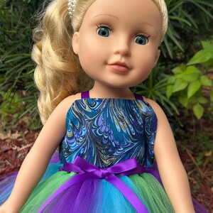 May include: A doll wearing a blue and green marbled print dress with a purple tulle skirt and a purple satin ribbon.