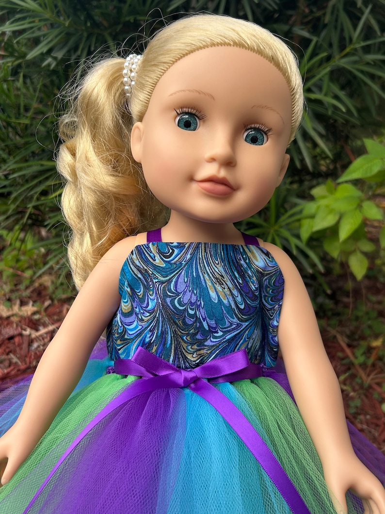 May include: A doll wearing a blue and green marbled print dress with a purple tulle skirt and a purple satin ribbon.