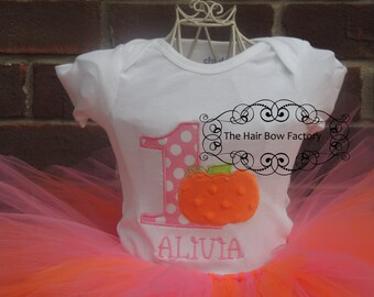 May include: A white onesie with a pink polka dot number one and an orange pumpkin. The onesie has the name "Alivia" embroidered on it. The onesie is paired with a pink and orange tulle tutu.