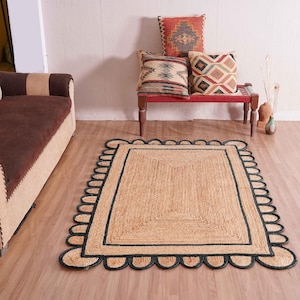 Puede incluir: Una alfombra rectangular de yute con un borde negro festoneado y un borde interior negro. La alfombra está colocada sobre un suelo de madera, con un sofá y cojines decorativos en el fondo. Dimensiones: aproximadamente 152 x 213 cm.