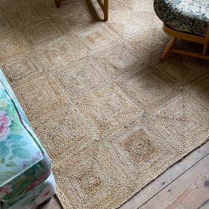 May include: A natural fiber rug with a square pattern. The rug is a light tan color and appears to be made of woven jute or similar material. The rug is placed on a wooden floor.