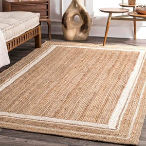 Puede incluir: Una alfombra rectangular de yute en color beige natural con un borde blanco. La alfombra tiene una textura tejida y está sobre un suelo de madera oscura. Es un artículo de decoración para el hogar.