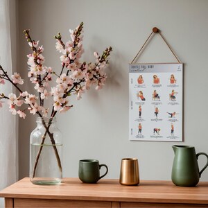 Puede incluir: Un p&oacute;ster con el texto "GENTLE FULL BODY workout" cuelga de una pared gris. El p&oacute;ster muestra ilustraciones de ejercicios. Un jarr&oacute;n de cristal transparente con ramas florecientes rosas, una taza verde, un jarr&oacute;n dorado y una jarra verde est&aacute;n sobre una mesa de madera.