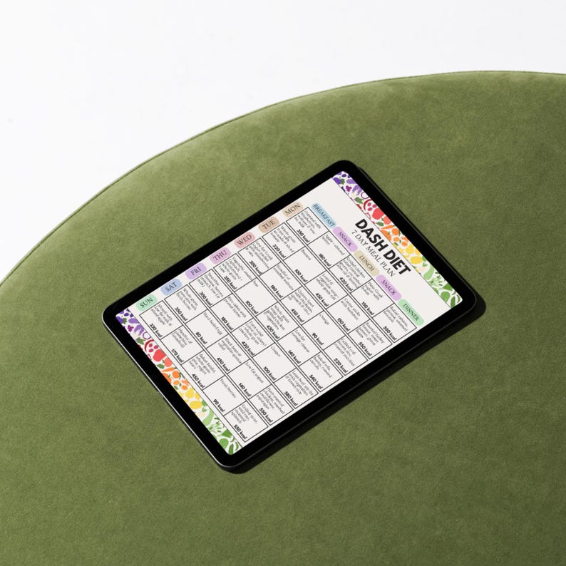 May include: A black tablet displaying a colorful DASH DIET 7 DAY MEAL PLAN. The plan is a grid with daily meal suggestions and calorie counts. The tablet is on a green surface.