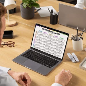 May include: A laptop displaying a "Mediterranean Diet 7 Day Meal Plan" on its screen. The laptop is open on a wooden desk, surrounded by office supplies, including pens, pencils, and a small potted plant. The screen shows a meal plan with various food options.