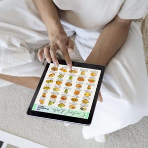 K&ouml;nnte beinhalten: Ein Tablet mit einer farbenfrohen Illustration von 30 Fr&uuml;hst&uuml;cksideen. Das Tablet wird von einer Person in wei&szlig;er Kleidung gehalten. Der Text "Cortisol Detox" und "30 Breakfast Ideas" ist auf dem Bildschirm zu sehen.