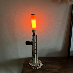 Puede incluir: Una lámpara beige y negra con una luz naranja y amarilla brillante. La lámpara está diseñada para parecerse a un rifle, con una base texturizada y una lámpara vertical. La lámpara está sobre una superficie de madera.