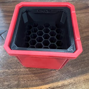 May include: A red and black square container with a honeycomb pattern inside. The top edge is red, and the interior is black. The text "6.5 CM" is visible on the top edge. The container sits on a wooden surface.
