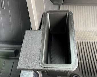May include: A black, rectangular car console organizer with a textured surface. The organizer has a deep, open compartment and is designed to fit into a vehicle's interior.