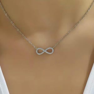 Puede incluir: Un collar plateado con un colgante en forma de símbolo de infinito incrustado con pequeñas piedras brillantes. La delicada cadena es visible sobre un fondo neutro, resaltando el diseño del colgante. Una pieza de joyería.
