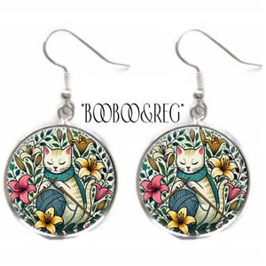 May include: A pair of silver dangle earrings with a round, colorful image of a white cat knitting yarn in a garden setting. The cat is wearing a blue scarf and the image is surrounded by a silver frame.