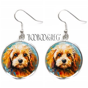 May include: A pair of silver-toned dangle earrings. Each earring features a round pendant with a colorful, artistic rendering of a dog's face. The dog has brown eyes and tan fur. The text "*BOOBOO&REG*" is above the earrings.