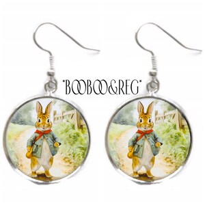 May include: A pair of silver earrings with round glass cabochons featuring a vintage illustration of a rabbit wearing a blue jacket and red scarf. The rabbit is walking down a path in a forest setting.