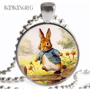 May include: A silver necklace with a round glass pendant featuring a cartoon illustration of a brown rabbit wearing a blue jacket, hopping over a wooden fence in a green field with flowers.