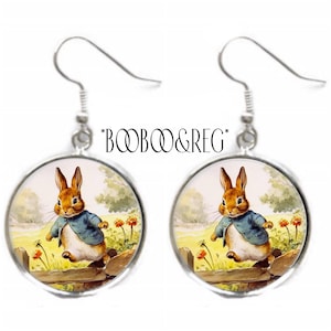 May include: A pair of silver dangle earrings featuring a round glass cabochon with a vintage illustration of a rabbit in a blue jacket hopping over a fence. The rabbit is brown with white fur on its belly and a pink nose. The background is a green field with yellow flowers.