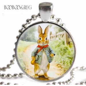 May include: A silver necklace with a round glass pendant featuring a vintage illustration of a brown rabbit wearing a blue jacket and red scarf. The rabbit is standing on a dirt path with green grass and a wooden fence in the background.