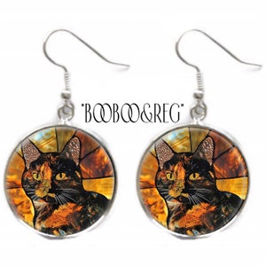 May include: A pair of silver dangle earrings featuring a round glass cabochon with a stained glass image of a tortoiseshell cat. The cat has yellow eyes and is set against a background of orange, yellow, and brown.