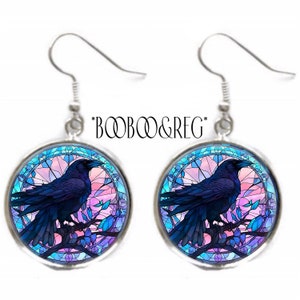 May include: A pair of silver-tone dangle earrings. Each earring features a round, glass-covered image of a dark blue raven perched on a branch, set against a stained-glass style background with pink and blue hues. The text "*BOOB&REG*" is above the earrings.
