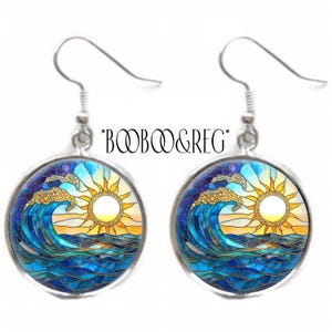 May include: A pair of round earrings with a stained glass design featuring a sun, ocean waves, and the text "*BOOB&GREG*". The earrings have silver-colored hooks and a blue, yellow, and gold color scheme.