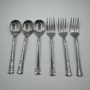 May include: Six-piece stainless steel flatware set. The set includes three spoons and three forks. Each piece has a simple, elegant design with a smooth handle and a decorative band near the base. The spoons have a rounded bowl, and the forks have four tines.