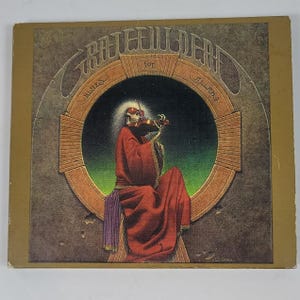 May include: A square vinyl record cover featuring the Grateful Dead album Blues for Allah. The artwork depicts a skeleton wearing red sunglasses and a long red robe, playing a violin. The figure is framed by a circular archway on a textured background.
