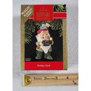 May include: Hallmark Keepsake ornament featuring a gnome holding a camera. The gnome wears a red hat, green boots, and a white beard. The box reads "MAGIC BLINKING LIGHT!" and "Holiday Flash". The ornament is in a gold and red box.
