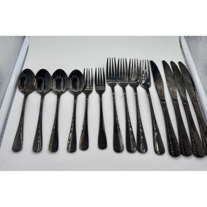 May include: A collection of silverware, including spoons, forks, and knives, arranged on a white tray. The silverware has a dark, metallic finish. The spoons are on the left, followed by forks and knives on the right. The utensils are for dining.