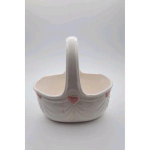 Puede incluir: Cesta de cerámica blanca con asa y detalles de corazones rosas. La cesta tiene forma curvada con un diseño festoneado alrededor del borde. El asa es arqueada y está unida a los lados de la cesta.