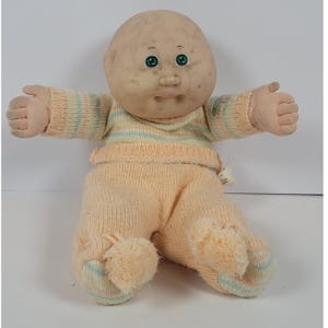 Puede incluir: Una muñeca calva con cara redonda pecosa y ojos verdes. La muñeca viste una camiseta de manga larga de punto color melocotón con finas rayas horizontales en azul claro y amarillo, combinada con pantalones de punto color melocotón a juego con pompones.
