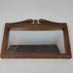 May include: A rectangular wall mirror featuring a medium-toned wooden frame. The top of the frame is adorned with a decorative, symmetrical wooden crest that includes a central cylindrical finial and curved, wing-like accents on each side.