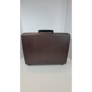 Puede incluir: Un maletín rectangular rígido de color marrón con una superficie texturizada y un asa de plástico negro centrada en el borde superior. El maletín presenta un diseño sólido y sencillo con esquinas redondeadas y una estructura robusta.