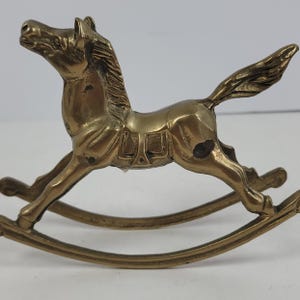 Könnte beinhalten: Eine Schaukelpferd-Figur aus Messing mit polierter Metalloberfläche, modellierter Mähne, detailliertem Sattel und fließendem Schweif. Das Pferd ist auf zwei geschwungenen Kufen montiert und zeigt ein klassisches Design mit sichtbarer Patina.