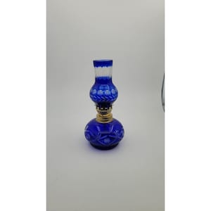 May include: A cobalt blue glass oil lamp featuring a rounded base and a matching chimney, both decorated with etched leaf and floral patterns. The lamp includes a metallic gold-colored burner assembly connecting the base to the chimney.