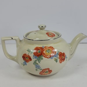 May include: A cream-coloured ceramic teapot featuring a floral pattern of orange poppies and small blue flowers. The teapot has a matching lid with a single orange poppy, a curved handle, a spout, and a silver-toned trim along the rim and lid.