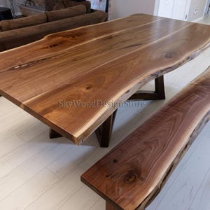 Puede incluir: Una gran mesa de comedor de madera con bordes naturales y un banco a juego. La mesa y el banco están hechos de madera marrón oscuro con vetas visibles y un borde natural. La mesa tiene una tapa rectangular y una base marrón oscuro. El banco tiene un diseño similar.