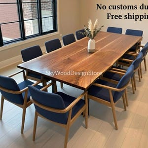 Puede incluir: Una gran mesa de comedor rectangular de madera con borde natural, rodeada de sillas tapizadas azules con marcos de madera. Un jarrón con tallos decorativos está sobre la mesa. La habitación tiene grandes ventanas.