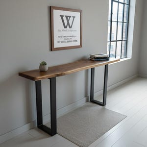 Pode incluir: Uma mesa de console de madeira com borda natural e pernas de metal preto. Uma impressão emoldurada com a letra "W" e o texto "Sky Wood Design Store" está pendurada na parede acima da mesa. Uma pequena planta em vaso e livros estão na mesa.