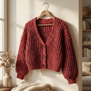 Op de afbeelding: Een bordeauxrode, grofgebreide cardigan met een V-hals en knoopsluiting, hangend aan een houten hanger. De geribbelde textuur en ballonmouwen geven een gezellige, stijlvolle uitstraling. De cardigan wordt tegen een neutrale muur getoond.