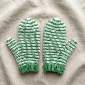May include: A pair of handmade mittens in a green and white striped pattern. The mittens are crocheted with a ribbed cuff and a rounded top. The background is a neutral tan fabric.