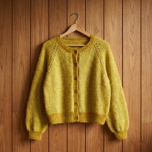 Può includere: Un cardigan giallo senape con scollo tondo e maniche lunghe, appeso a una gruccia di legno. Il maglione a maglia presenta una parte anteriore con bottoni e un design strutturato. Lo sfondo è un muro di legno.