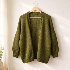 Può includere: Cardigan lavorato a maglia verde oliva con maniche lunghe e vestibilità ampia, appeso a una gruccia di legno. Il maglione aperto presenta un motivo a maglia strutturato e un design classico e confortevole. Il cardigan è presentato su uno sfondo neutro.