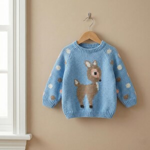 May include: A light blue knit sweater with a deer design on the front. The sweater has a crew neck and long sleeves with polka dots. The deer is brown with a white chest and pink accents. The sweater is hanging on a wooden hanger.