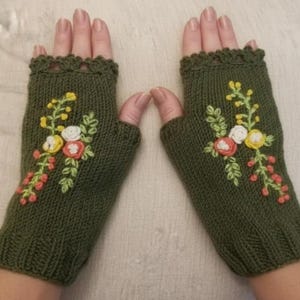 Puede incluir: Un par de guantes sin dedos de color verde oliva con bordados florales. Los guantes tienen un borde festoneado y están adornados con flores bordadas en amarillo, blanco y coral. Los guantes de punto se muestran sobre un fondo neutro.