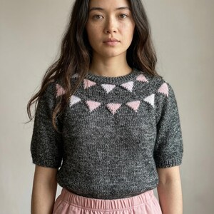 May include: A gray knit sweater with short sleeves and a crew neck. The sweater features a pattern of pink and white triangle shapes across the upper chest. The model is wearing pink pants.