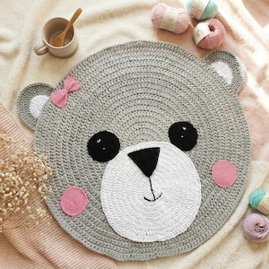 May include: A handmade, gray crochet bear rug with a white face, black eyes, and a black nose. The rug features pink accents, including a bow and cheek circles. The rug is round and has bear ears. Yarn balls and a mug are in the background.