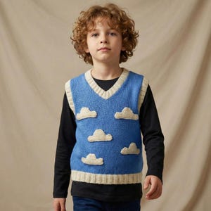 Puede incluir: Un chaleco azul tejido con cuello en V y ribete color crema. El chaleco presenta aplicaciones de nubes blancas. La persona lleva debajo una camiseta negra de manga larga. Es una prenda de vestir infantil.