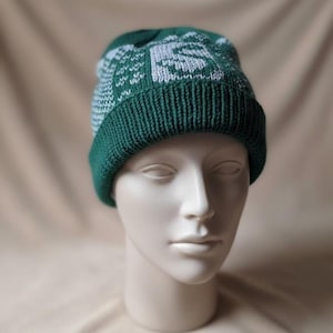 May include: A green and white knit beanie hat. The hat features a folded brim and a design on the crown. The hat is displayed on a mannequin head against a neutral background. The hat is made of a soft, knitted material.