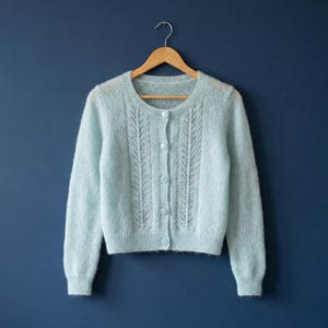 May include: A pale blue, long-sleeved cardigan with a round neck and button closure. The knitted garment has an intricate, openwork pattern down the front. The cardigan is displayed on a wooden hanger against a dark blue backdrop.